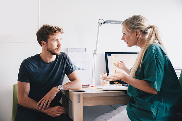 En kvinnling sjuksköterska talar med en manlig patient vid ett skrivbord.